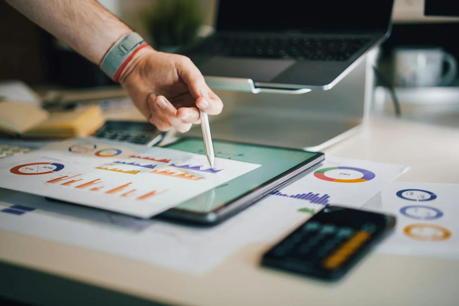 A hand holding a stylus over a tablet, among printed charts and a calculator